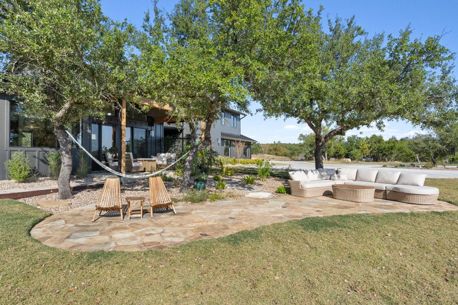 Expansive flagstone patio with mature shade trees, a manicured lawn, and a modern exterior featuring large glass panels