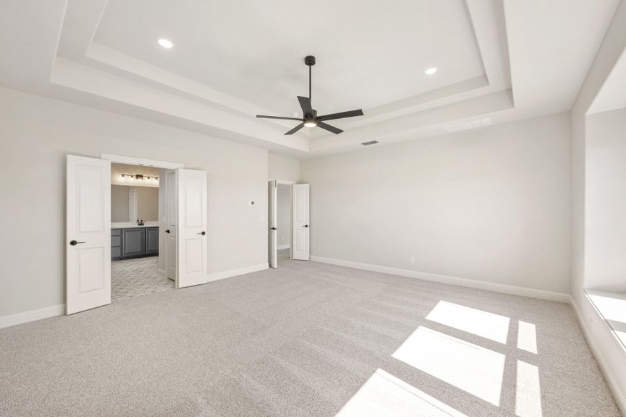 Large master bedroom with tray ceiling, ceiling fan, carpet flooring, and open doors to ensuite bathroom and closet. Large master bedroom with tray ceiling, ceiling fan, carpet flooring, and open doors to ensuite bathroom and closet.