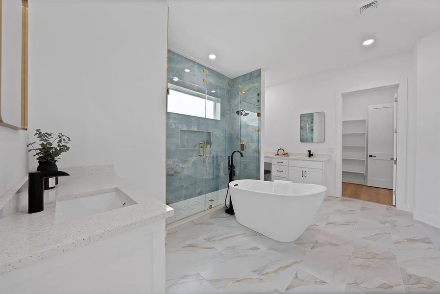 Freestanding soaking tub with a floor-mounted matte black faucet, a walk-in shower featuring a tiled accent wall and glass enclosure, and dual white vanities with integrated sinks and quartz countertops