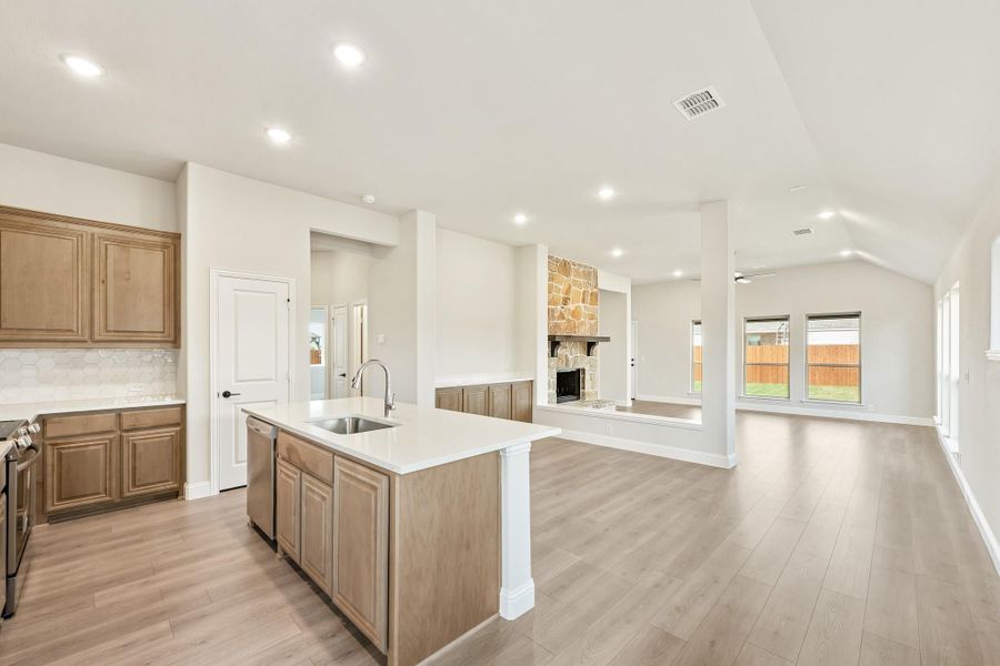 Open-concept kitchen with center island, wood cabinets, and stone fireplace visible in living area beyond Open-concept kitchen with center island, wood cabinets, and stone fireplace visible in living area beyond