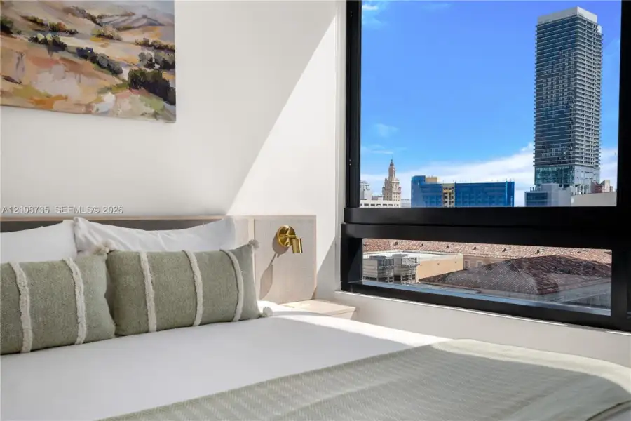 Floor-to-ceiling windows featuring urban skyline scenery, built-in light-toned headboard with integrated gold-tone sconce Floor-to-ceiling windows featuring urban skyline scenery, built-in light-toned headboard with integrated gold-tone sconce