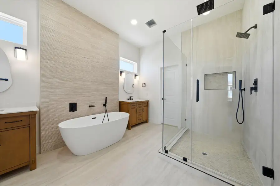 This modern bathroom features a spacious glass-enclosed shower, a sleek freestanding tub, and dual vanities with wood cabinetry. The neutral tones and large windows create a bright, airy atmosphere.