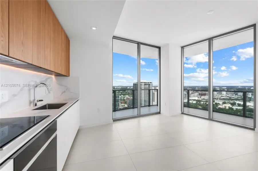 Modern kitchen featuring slab-front wood cabinetry, white stone-finish countertops, full-height stone-finish backsplash, built-in cooktop, and floor-to-ceiling windows with balcony access