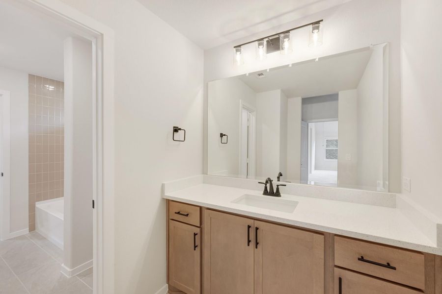Bathroom vanity with wood cabinets, white countertop, large mirror, and soaking tub visible in background