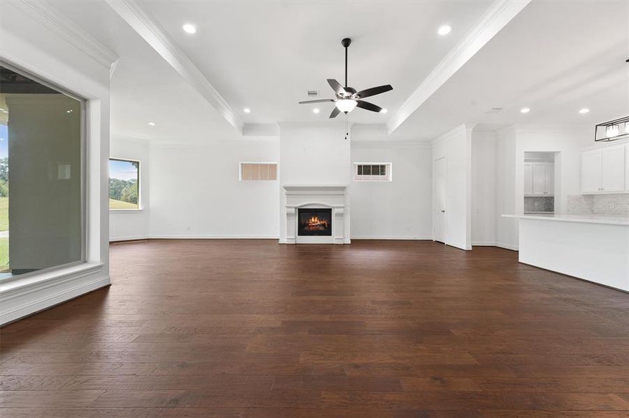 This is a spacious, well-lit living area with hardwood floors, a fireplace, and an open layout connecting to the kitchen. It features large windows, a tray ceiling with recessed lighting, and a modern ceiling fan.