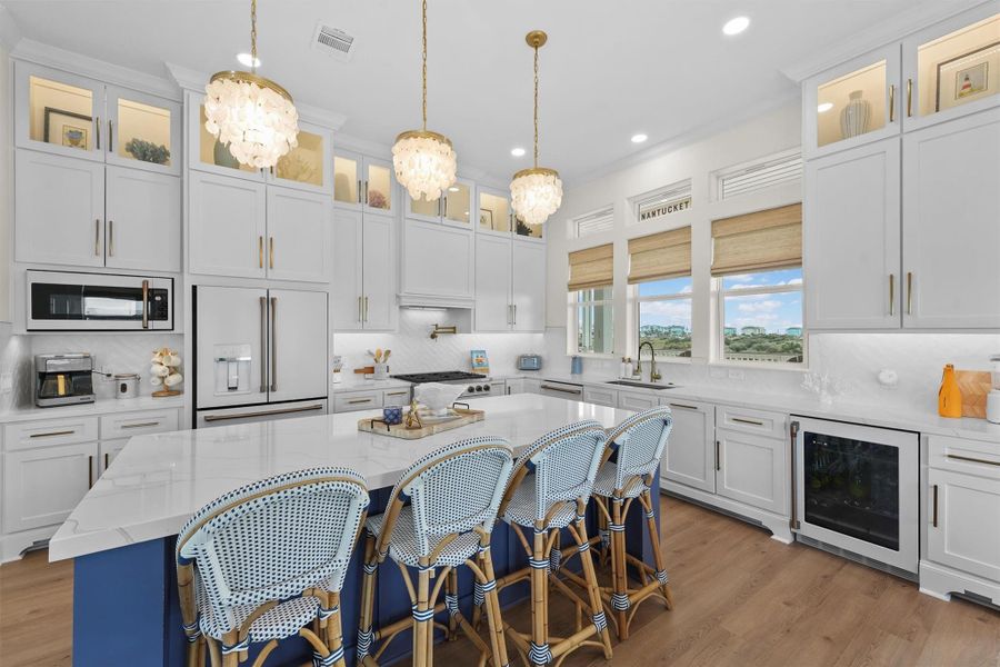 This kitchen features a bright, modern design with upgraded Cafe appliances, Cafe wine refrigerator, white floor to ceiling cabinetry and quartz countertops, accented by gold hardware. A large island with 4 barstools offers ample seating. Elegant pendant lights hang above, and multiple windows provide natural light and scenic views accented. A built-in wine cooler and upgraded Cafe  appliances add convenience and style.