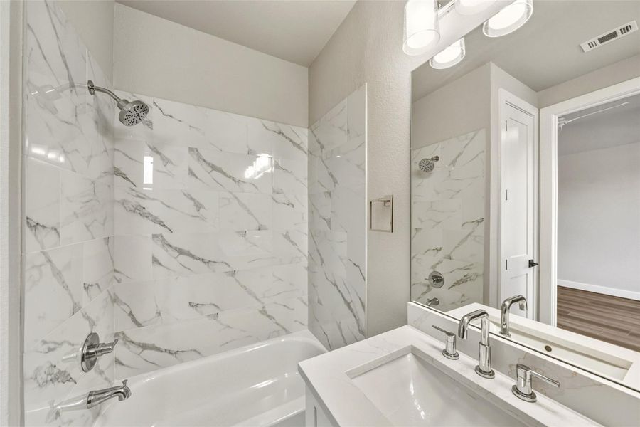 This bathroom features a sleek design with a bathtub and shower combo, adorned with elegant marble-style tiles. The modern vanity includes a white countertop, a stylish sink, and contemporary fixtures, complemented by overhead lighting.