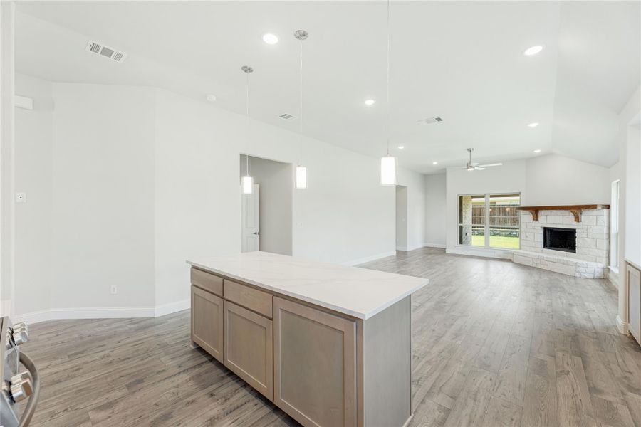 Open-concept kitchen and living area with center island, pendant lights, wood floors, and stone fireplace Open-concept kitchen and living area with center island, pendant lights, wood floors, and stone fireplace