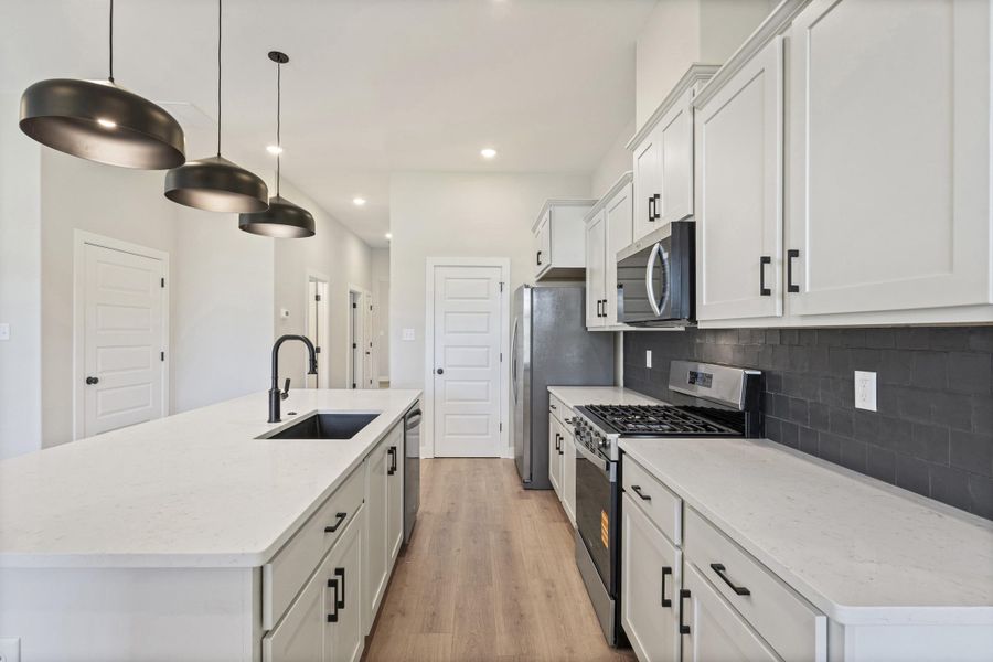 Kitchen featuring an expansive island with a matte black faucet and undermount sink, pendant lighting, light wood-finish flooring, stainless steel appliances, and white cabinetry with black hardware Kitchen featuring an expansive island with a matte black faucet and undermount sink, pendant lighting, light wood-finish flooring, stainless steel appliances, and white cabinetry with black hardware
