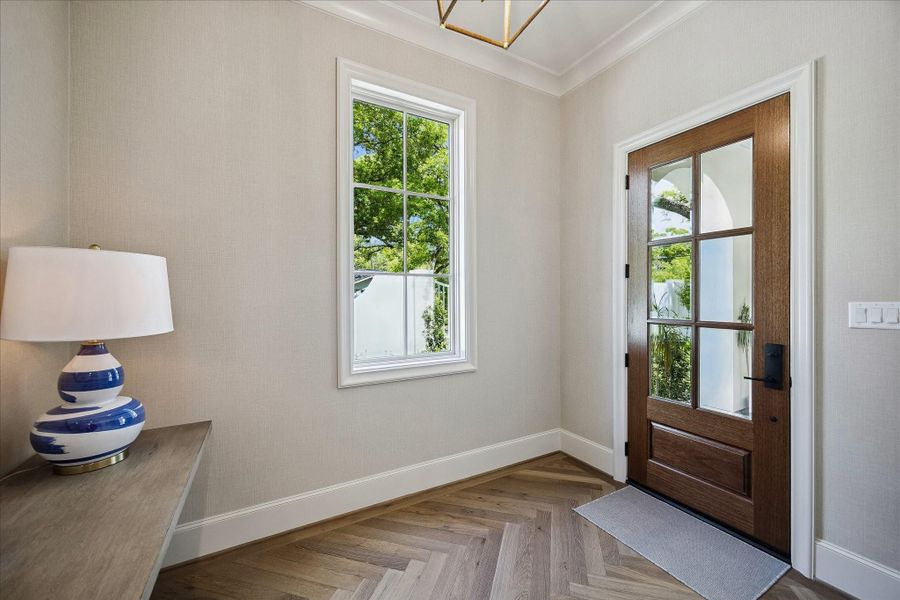 Additional view of the front foyer showcasing beautiful herringbone hardwood floors, detailed mill work and elegant glass-paneled wood-stained entry door. Natural light pours through the window, highlighting the clean lines and refined finishes.