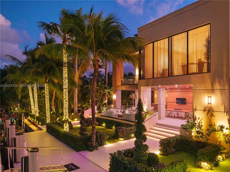 Dusk view of waterfront side yard with string-lit royal palms, manicured hedges, illuminated walkway, dock access, covered loggia with outdoor dining and TV, glass-railed terrace, and glowing two-story floor-to-ceiling glass facade.
