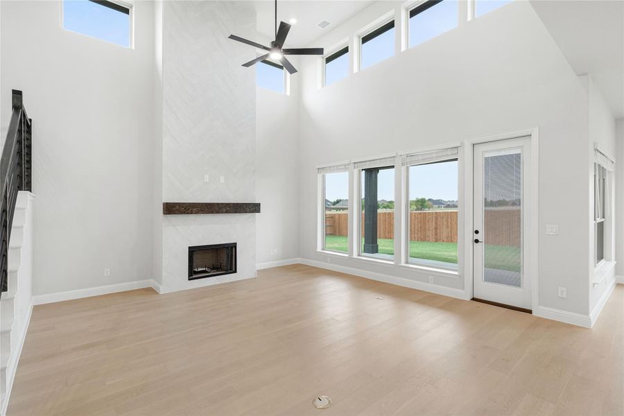 Open living room with two-story ceiling, tile fireplace, ceiling fan, and large windows with backyard view