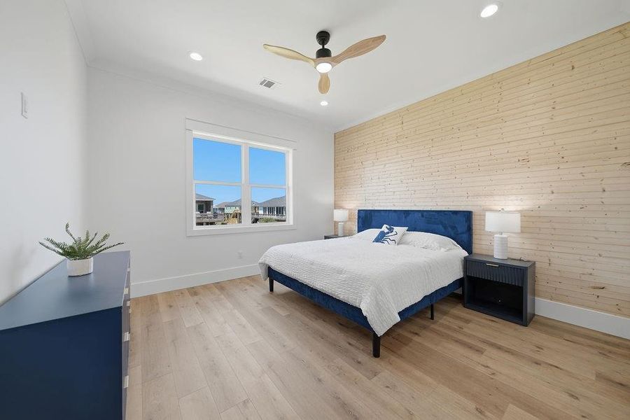 This bright bedroom features a wooden accent wall, a large window with natural light, and modern decor with a blue bed frame and matching dresser. The light wood flooring and ceiling fan add a warm touch.