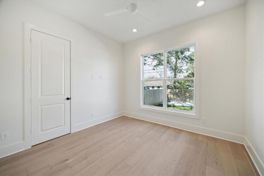 A first-floor bedroom offers versatility and comfort with its spacious layout and natural light. Large windows frame serene backyard views, filling the room with warmth and brightness. Crisp white walls and light wood flooring create a fresh, neutral backdrop ready for personalization. Recessed lighting and a modern ceiling fan enhance the functionality and style of the space. Whether used as a guest room, home office, or flex space, this room offers convenience and privacy on the main level.