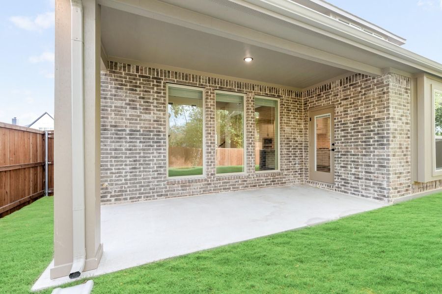 Covered back patio with concrete floor, brick exterior, sliding glass door, and green lawn with wood privacy fence.