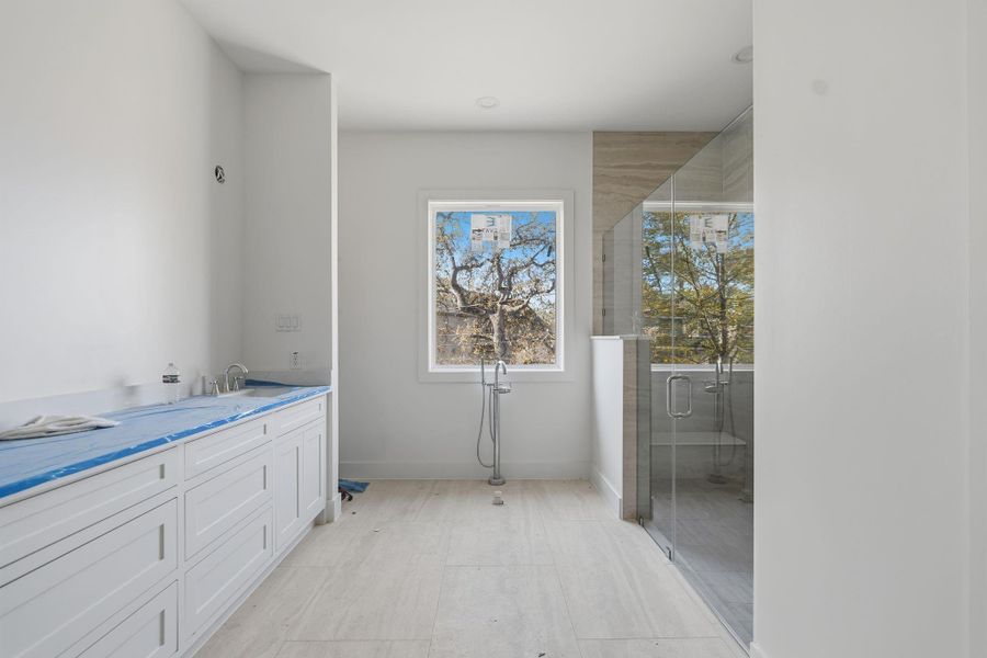 This view shows the layout of the primary bath, with a freestanding soaking tub to be installed beneath the window, a glass walk in shower to the right, and dual vanities along the opposite wall.
