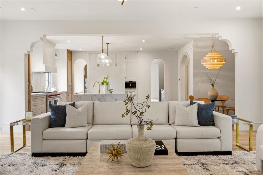 Living area featuring a sectional sofa, wood-finish flooring, recessed lighting, and arched doorways opening to a kitchen with a waterfall island and pendant lighting Living area featuring a sectional sofa, wood-finish flooring, recessed lighting, and arched doorways opening to a kitchen with a waterfall island and pendant lighting