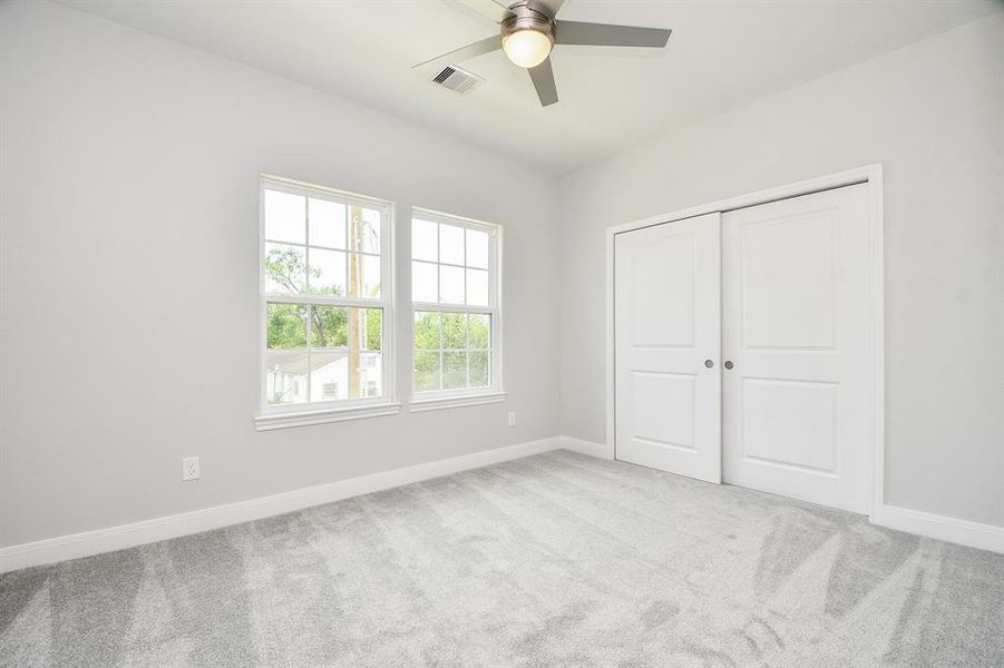 This is a bright, secondary bedroom featuring plush carpeting, two large windows providing natural light, a modern ceiling fan, and a double-door closet. The walls are a neutral gray, offering a versatile backdrop for any decor.