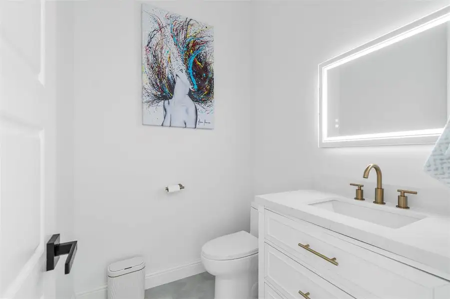 Bathroom featuring a white vanity with gold-finish hardware, an integrated sink, a gold-finish faucet, and an illuminated mirror