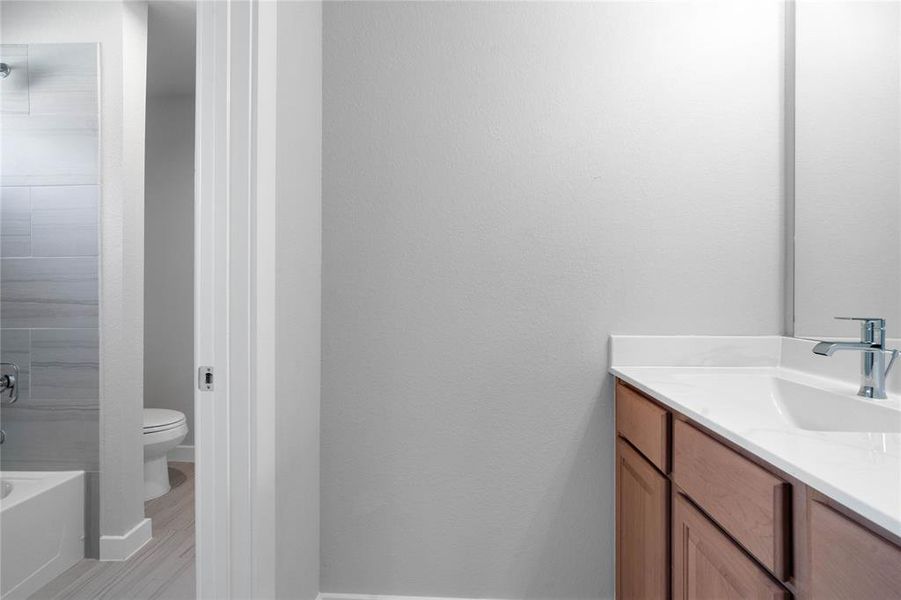 The Jack and Jill bath features tile flooring, bath/shower combo with tile surround, stained wood cabinets, beautiful light countertops, mirror, dark, sleek fixtures and modern finishes.