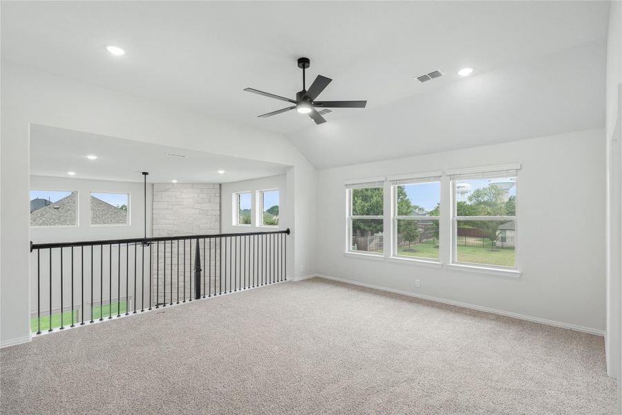 Open upstairs loft with carpet, black ceiling fan, metal railing, and large windows overlooking backyard. Open upstairs loft with carpet, black ceiling fan, metal railing, and large windows overlooking backyard.
