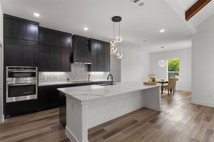 Kitchen with tasteful backsplash, dark cabinets, light stone countertops, decorative light fixtures, and custom exhaust hood