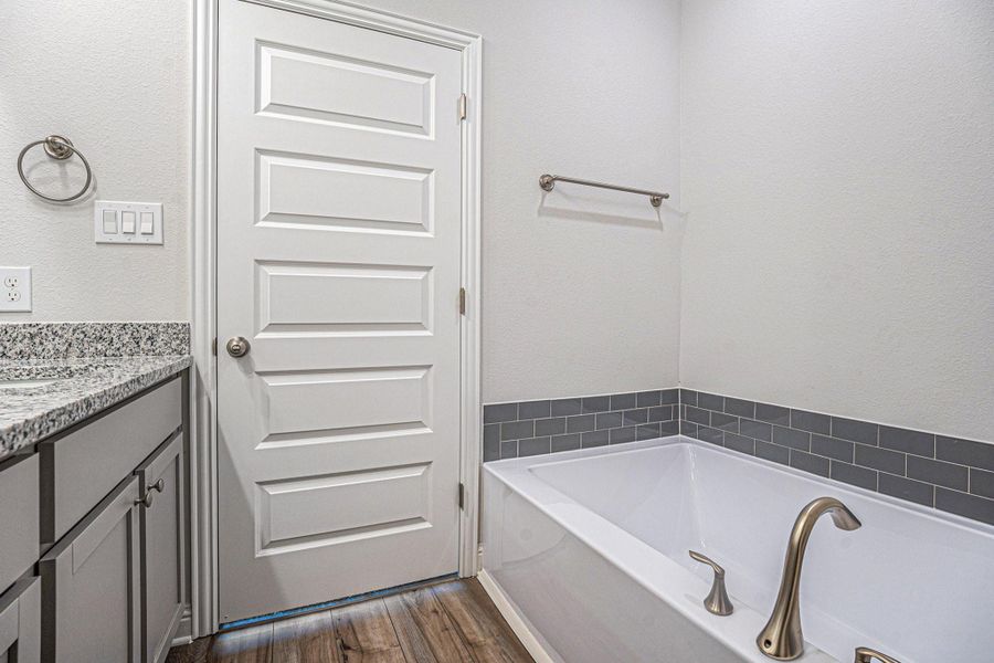 This primary bathroom features a modern design with a granite countertop, a sleek under-mount sink, and a spacious soaking bathtub with a brushed nickel faucet. The room is accented by gray tile and luxury vinyl plank flooring.