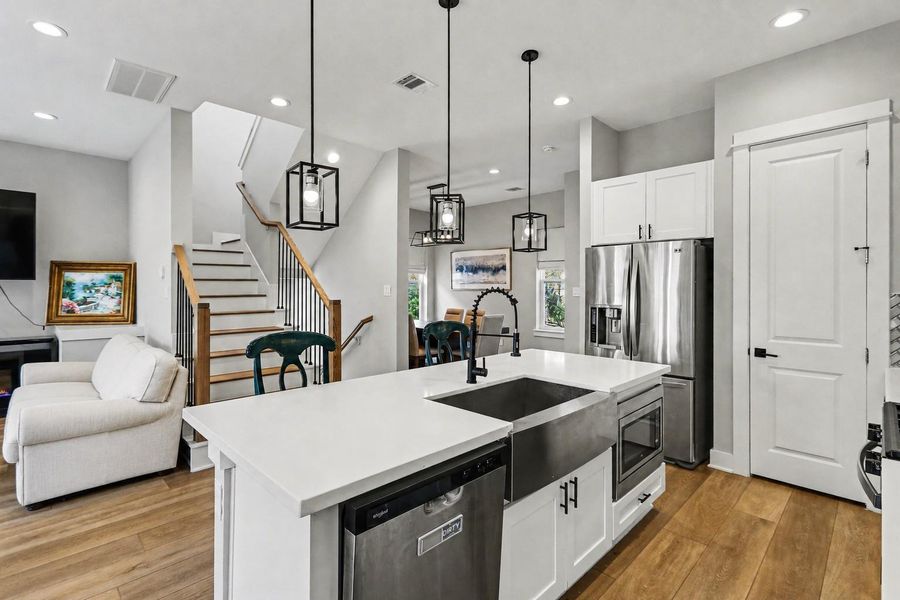 This modern kitchen features a spacious island with a sink and dishwasher, stainless steel appliances, and pendant lighting. It opens into a bright living area with hardwood floors and a staircase, creating an inviting and functional space.