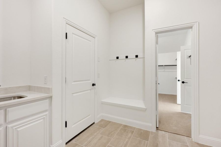 Mudroom with built-in bench, wall hooks, white cabinets with sink, and tile floors leading to a closet Mudroom with built-in bench, wall hooks, white cabinets with sink, and tile floors leading to a closet