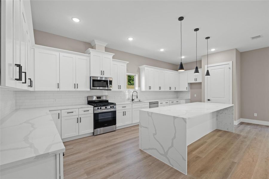 Modern kitchen featuring white cabinetry, sleek countertops, and a spacious island with pendant lighting. Stainless steel appliances complement the bright, open layout and wood flooring adds warmth. Ideal for entertaining and culinary endeavors.