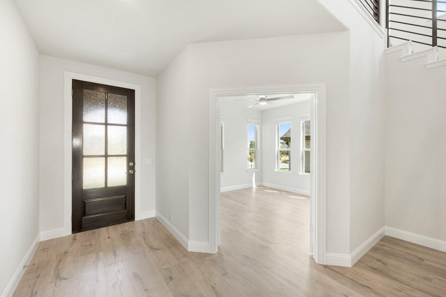 Entryway with dark wood glass-panel front door, light hardwood floors, and open view into adjacent room with ceiling fan.