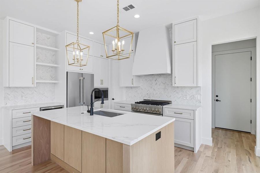 Kitchen with an island with sink, light stone counters, hanging light fixtures, light wood finished floors, and open shelves Kitchen with an island with sink, light stone counters, hanging light fixtures, light wood finished floors, and open shelves