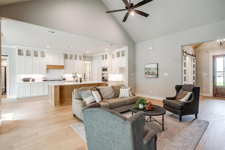 Living area with a barn door, visible vents, light wood-style flooring, high vaulted ceiling, and ceiling fan Living area with a barn door, visible vents, light wood-style flooring, high vaulted ceiling, and ceiling fan