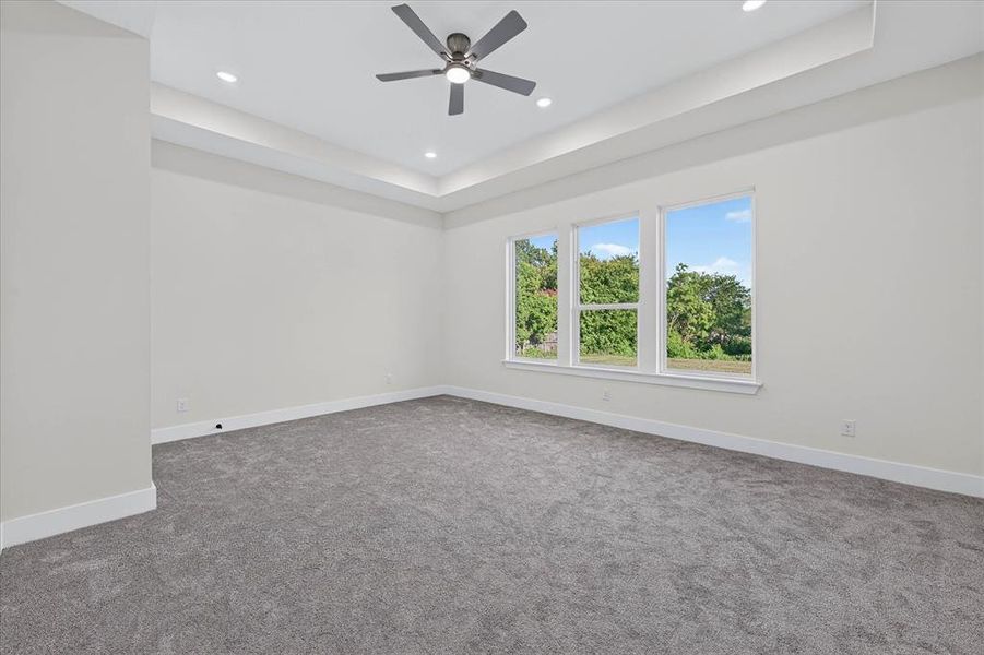 Spacious room featuring triple windows, tray ceiling with recessed lighting, gray carpet flooring, a ceiling fan, and white baseboards Spacious room featuring triple windows, tray ceiling with recessed lighting, gray carpet flooring, a ceiling fan, and white baseboards