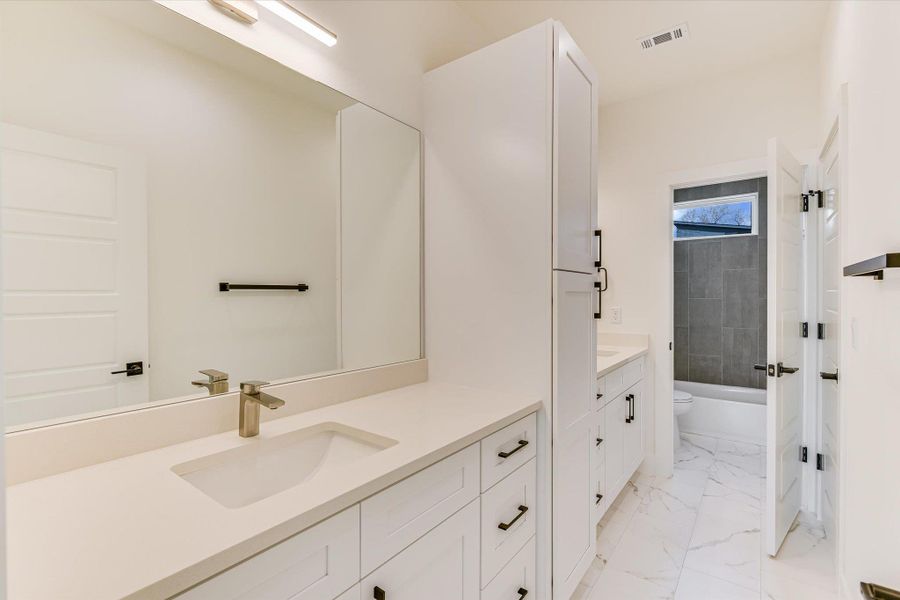 The ultimate in functional design, this Jack 'n Jill bathroom offers plenty of storage with a dedicated floor-to-ceiling linen cabinet. The high-contrast aesthetic is defined by white quartz countertops paired with bold black hardware and marble-style flooring that flows into the private shower area.