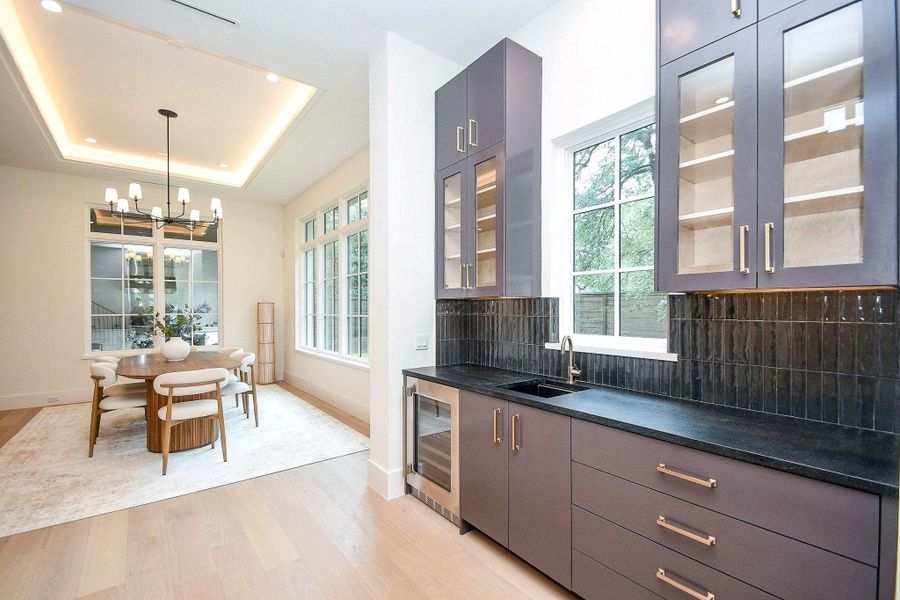 This photo showcases a modern dining area with an adjacent sleek bar space. It features a spacious dining table under a stylish chandelier, large windows providing natural light, and elegant cabinetry with glass doors. The room has a contemporary feel with light wood flooring and a neutral color palette.
