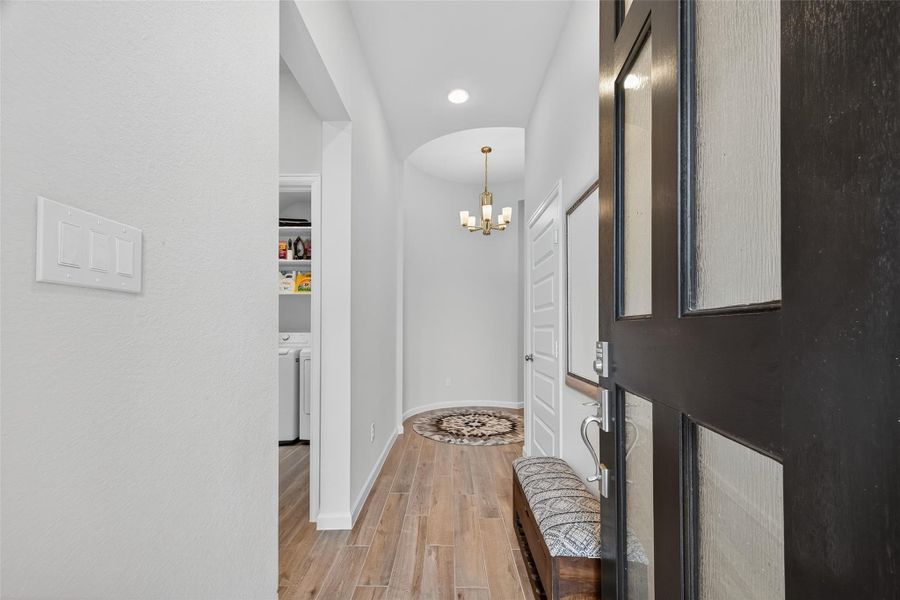 This inviting entryway features hardwood flooring, a modern chandelier, and a cozy bench. The open door reveals a glimpse of a laundry area to the left, leading into a bright, welcoming hallway opening up to a foyer with a charming rotunda. This inviting entryway features hardwood flooring, a modern chandelier, and a cozy bench. The open door reveals a glimpse of a laundry area to the left, leading into a bright, welcoming hallway opening up to a foyer with a charming rotunda.