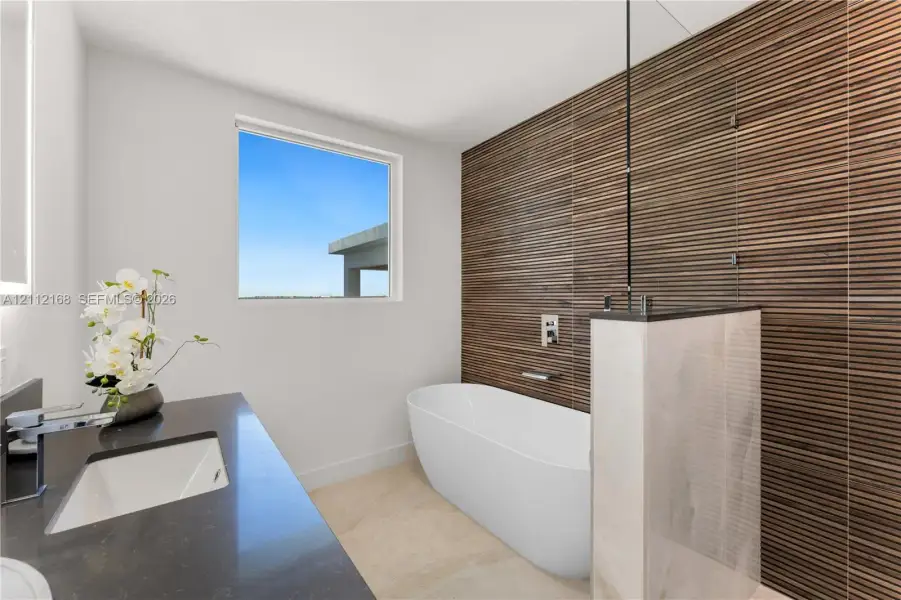 Freestanding soaking tub featuring a wall-mounted faucet, horizontal slatted wood-texture accent wall, glass partition, stone-finish vanity countertop, and rectangular undermount sink Freestanding soaking tub featuring a wall-mounted faucet, horizontal slatted wood-texture accent wall, glass partition, stone-finish vanity countertop, and rectangular undermount sink