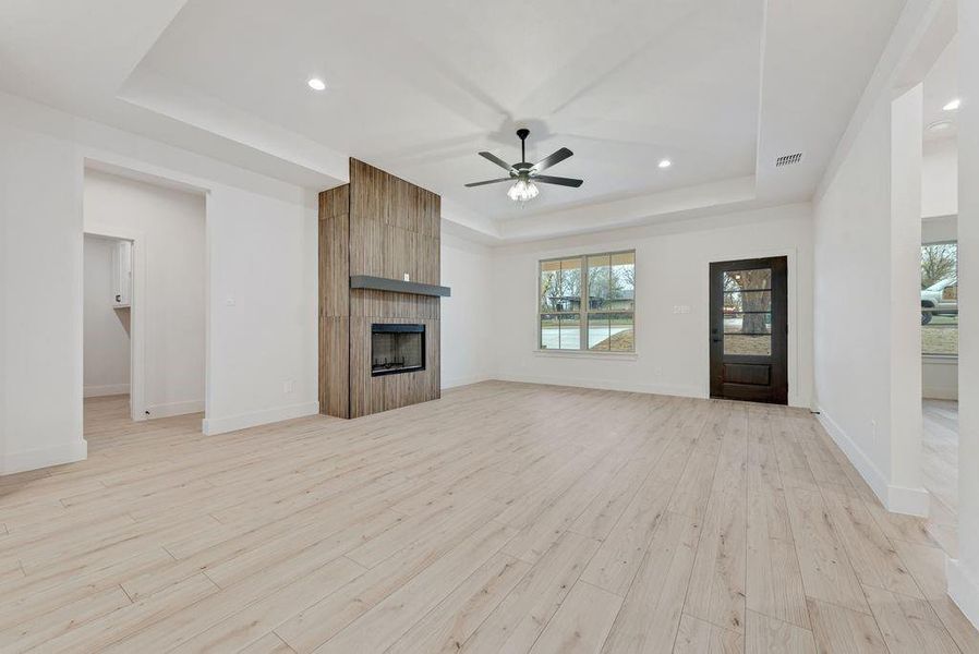 Unfurnished living room featuring a tray ceiling, light wood-style floors, a large fireplace, recessed lighting, and a ceiling fan