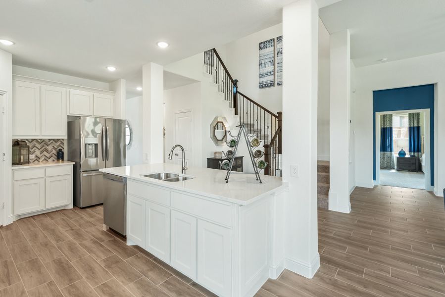 Kitchen with white island and sink, stainless steel appliances, tile backsplash, and wood-look floors open to staircase