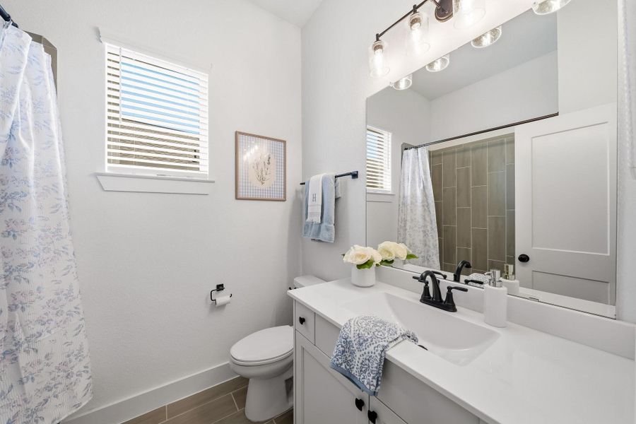The secondary bath has a window with blinds and features rich wood tile flooring, a sleek quartz-topped vanity, and a decorative light fixture that adds a finishing touch to this stylish and functional space.