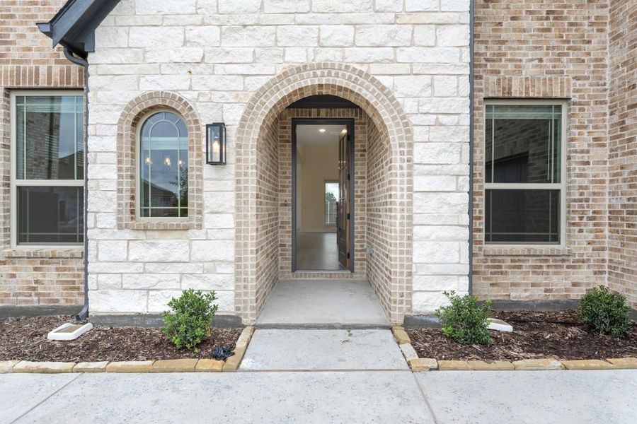 Home entrance with brick arched doorway, stone and brick exterior, wall sconce, and small landscaped shrubs flanking the entry.