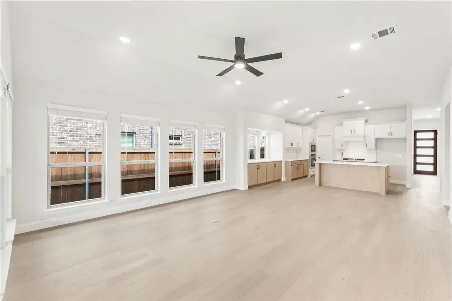 Open living area with light wood floors, ceiling fan, and kitchen with white cabinets and island in background