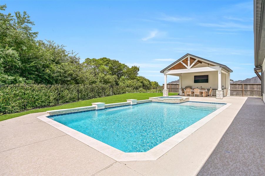 Looking across the sparkling Aqua Sport pool, the raised spa and covered outdoor kitchen pavilion create a resort-like focal point, while the tree-lined rear boundary provides a lush backdrop and added privacy.