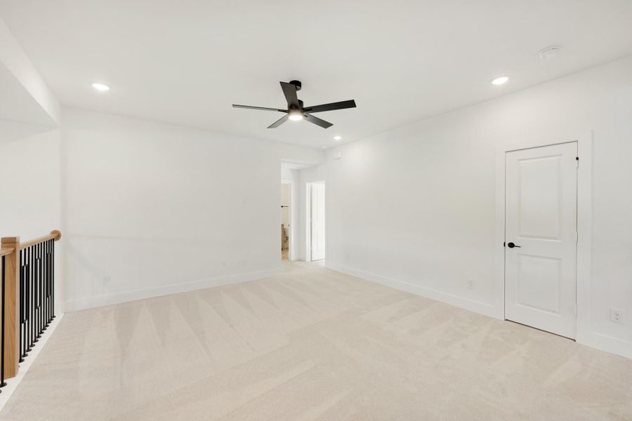Empty room with beige carpet, black ceiling fan, white walls, and staircase railing visible