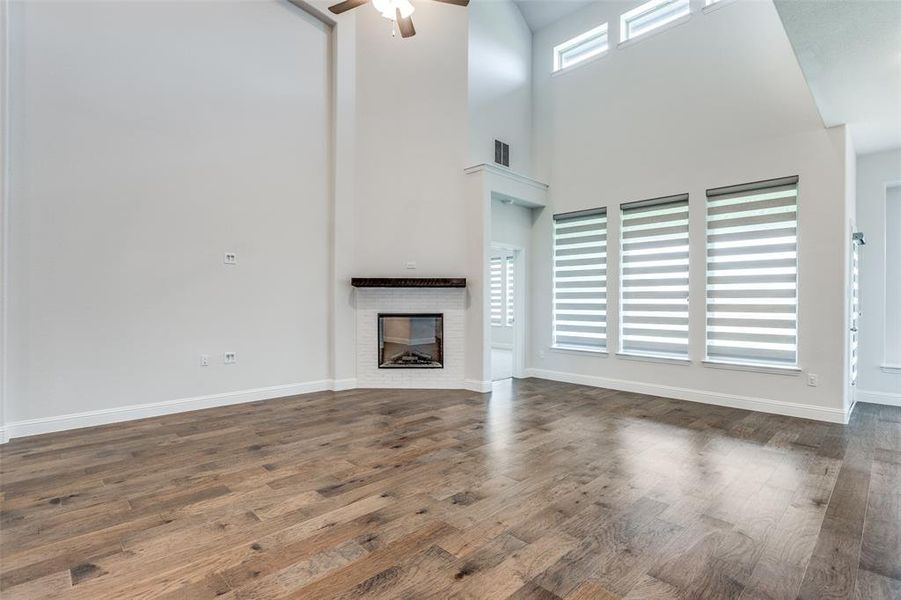 Unfurnished living room with a ceiling fan, wood finished floors, a brick fireplace, baseboards, and a high ceiling