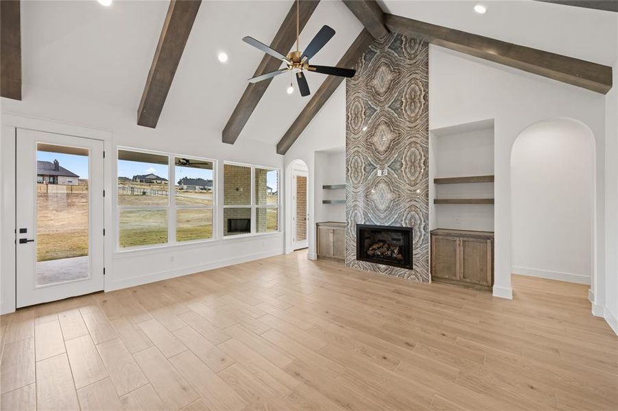 Vaulted ceilings with exposed wood beams, a striking fireplace with a full-height decorative surround, light wood-finish flooring, recessed lighting, and built-in shelving with cabinetry