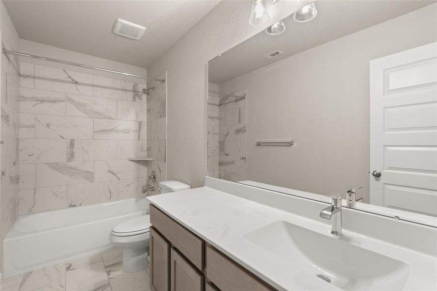 This bathroom features a modern design with a spacious vanity, sleek countertop, and a large mirror. It includes a bathtub with elegant marble-style tile, a toilet, and stylish fixtures. The neutral color palette adds a clean and inviting feel.