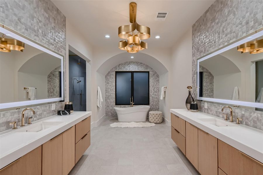 The bathroom features dual vanities with light wood cabinetry, illuminated mirrors, and a freestanding bathtub set beneath an arched alcove with tiled walls