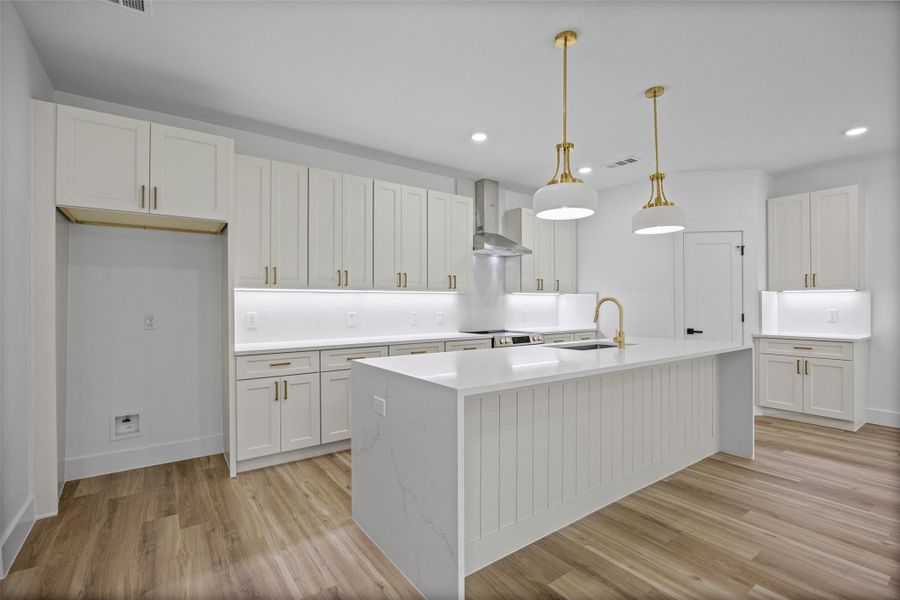 Modern kitchen featuring a large central island with paneling, white cabinetry with gold-tone hardware, quartz countertops and backsplash, wood-finish flooring, and recessed lighting Modern kitchen featuring a large central island with paneling, white cabinetry with gold-tone hardware, quartz countertops and backsplash, wood-finish flooring, and recessed lighting